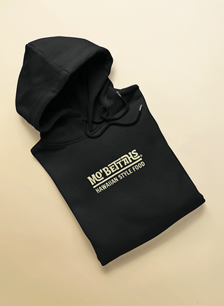 Black hoodie with 'Mo' Bettas Hawaiian Style Food' text on a sand background