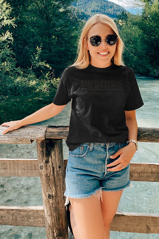 Young adult woman standing on bridge, over river, wearing black t-shirt with Mo' Bettahs dark grey logo on front.