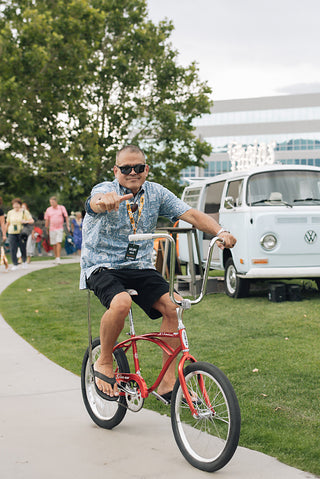 Co-founder, Kalani Mack, riding a red Schwinn Sting Ray, high handle bar bicycle, waving 'Hang Loose'.