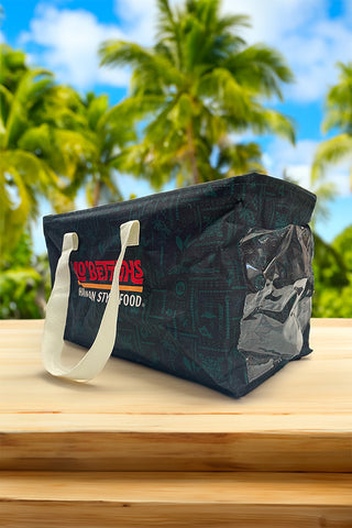 Mo' Bettahs Utility Tote on wood counter top, in tropical setting. Showing Mo' Bettahs Hawaiian Style Food logo, carrying straps, and side vinyl pocket.