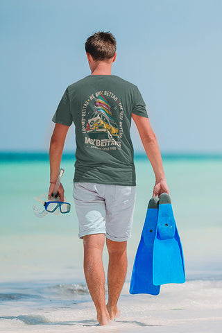 Back view of man carrying snorkling mask and fins, walking along beach, wearing sage green T-shirt with Mo' Bettahs VW bus stacked with surf boards graphic on the back.